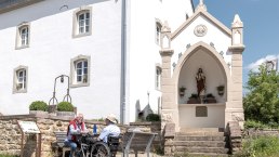 Zwei Männer, einer im Rollstuhl, sitzen auf einer Bank vor einem weißen Gebäude mit einer religiösen Statue in einer Nische. Sonniger Tag., © Naturpark Südeifel, Thomas Urbany