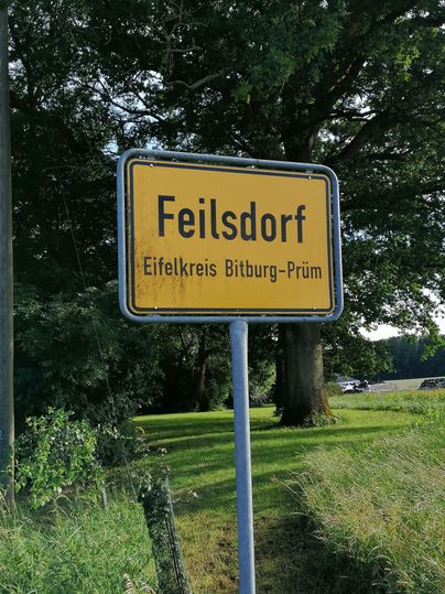 Een plaatsnaambord met de tekst "Feilsdorf" in de Eifelkreis Bitburg-Prüm. Omgeven door bomen en gras.