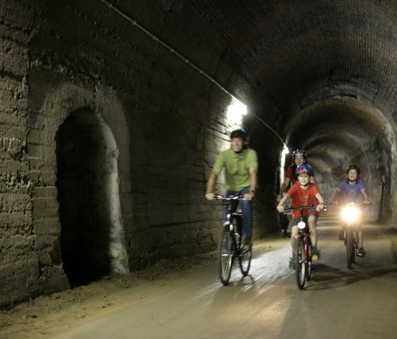 Drei Radfahrer, darunter ein Kind, fahren durch einen beleuchteten Tunnel. Der Tunnel ist Teil des Eifel-Ardennen-Radwegs, ehemals eine Bahnstrecke., &copy; Eifel Tourismus GmbH/intention