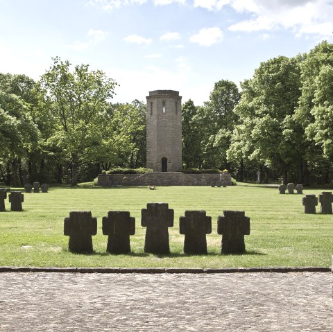 Ein Kriegsdenkmal mit Steinkreuzen auf einer Wiese, ein Turm im Hintergrund, umgeben von gr&uuml;nen B&auml;umen unter blauem Himmel., &copy; TI Bitburger Land