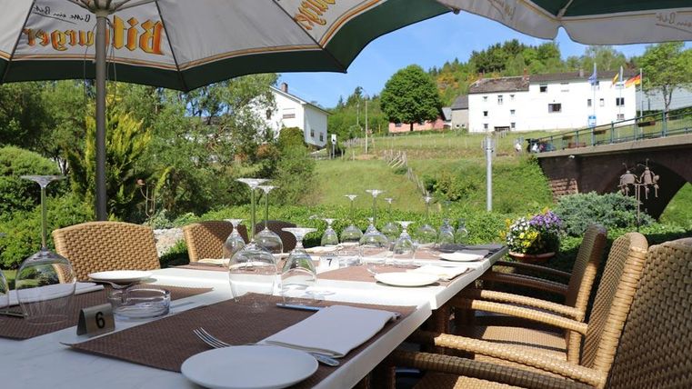 Une table accueillante en plein air avec des chaises en rotin, dressée avec des verres et de la vaisselle. À l'arrière-plan, on peut voir des collines verdoyantes et un bâtiment charmant.