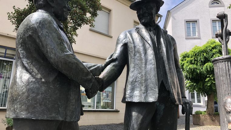 Eine bronze Statue von zwei Männern, die sich die Hände schütteln. Im Hintergrund sind alte Gebäude und Bäume sichtbar.