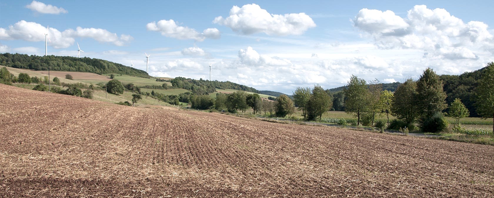 Wide landscape with fields, trees and wind turbines in the background. The sky is blue with white clouds., &copy; V. Teuschler