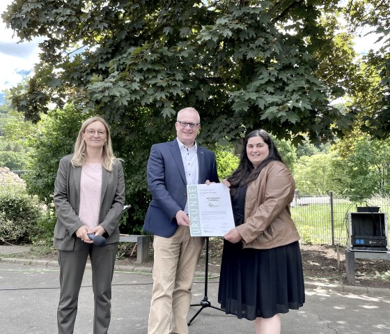 Michael Holstein (B&uuml;rgermeister Verbandgemeinde Trier-Land) und Daniela Torgau (Gesch&auml;ftsf&uuml;hrerin Naturpark S&uuml;deifel, links) &uuml;bergeben die Urkunde an Claudia Barzen (Schulleiterin Grundschule Ralingen)., &copy; Naturpark S&uuml;deifel/Ansgar Dondelinger