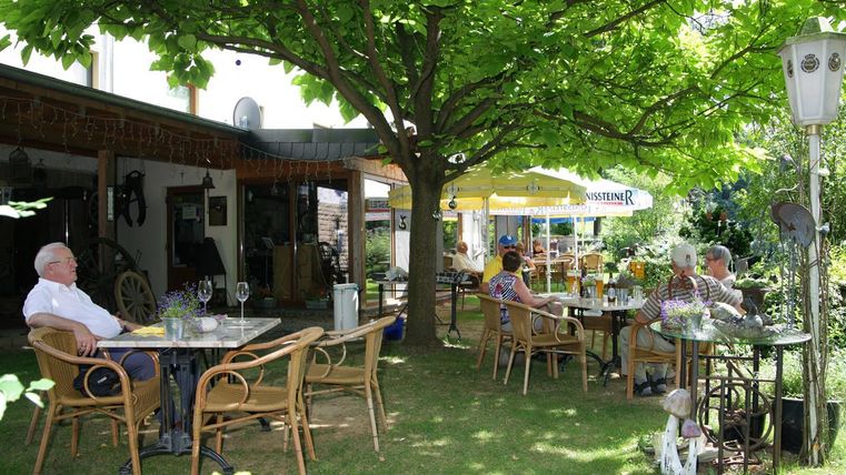 Un café confortable en plein air avec plusieurs tables et chaises sous un grand arbre. Les invités profitent de l'ambiance détendue dans un jardin verdoyant.