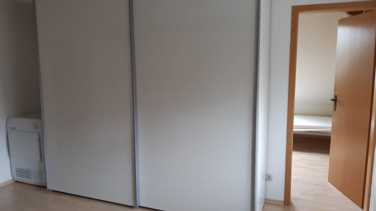 Een eenvoudige kamer met een grote, witte kast en een open deurkozijn. De vloer is gemaakt van licht hout en er is een airconditioningeheid in de hoek.