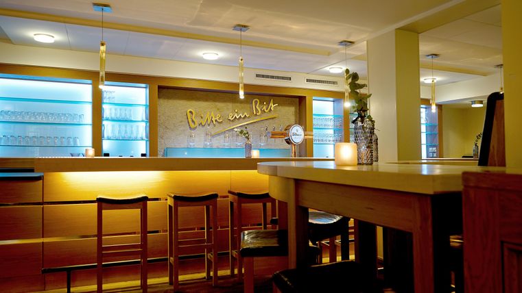 A cozy restaurant with an elegant bar and warm lighting. The walls are decorated in light colors, and there are some plants for an inviting atmosphere.