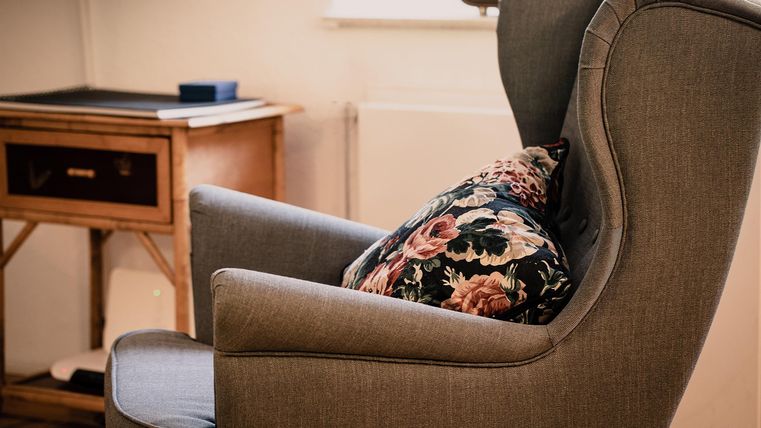 Ein bequemer Sessel mit einem bunten Kissen steht in einem gemütlichen Raum. Im Hintergrund sind ein kleiner Tisch und ein Fenster sichtbar.