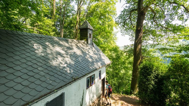 Ein Wanderer betritt eine kleine Einsiedelei im Wald mit Schieferdach und Glockenturm.