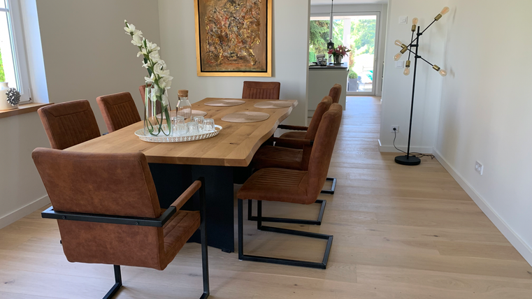 Een moderne eetruimte met een grote houten werktafel en stijlvolle stoelen. In de hoek staat een decoratieve vloerlamp en een groot raam dat veel licht binnenlaat.