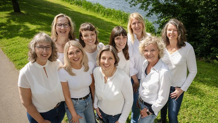 Eine Gruppe von Frauen in formeller Kleidung steht vor einem Springbrunnen in einem Park. Im Hintergrund sind mehrere Flaggen und gebaute Strukturen sichtbar.
