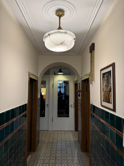 Un couloir lumineux avec un plafonnier décoratif et des carreaux colorés sur les murs. Au bout du couloir, des portes sont visibles, menant à d'autres pièces.