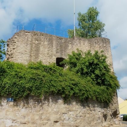Eine alte, teilweise zerstörte Burg ist von grünem Gestrüpp umgeben. Der Himmel ist klar mit einigen Wolken.