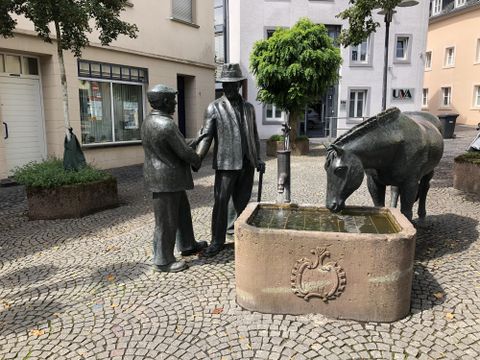 Eine Bronze-Skulptur von zwei Menschen, die sich die Hand schütteln, steht neben einem Pferd und einem Brunnen. Der Platz ist von einer steinernen Pflasterung umgeben.