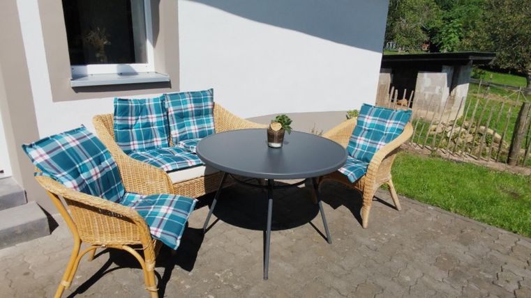 Een gezellige zitruimte buiten met een ronde tafel en drie rotanstoelen. De stoelen zijn bekleed met blauwe en groene kussens.