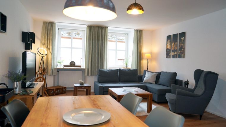 Een gezellige woonkamer met een moderne bank en een eetgedeelte. Grote ramen laten veel daglicht binnenkomen en geven de ruimte een uitnodigende sfeer.