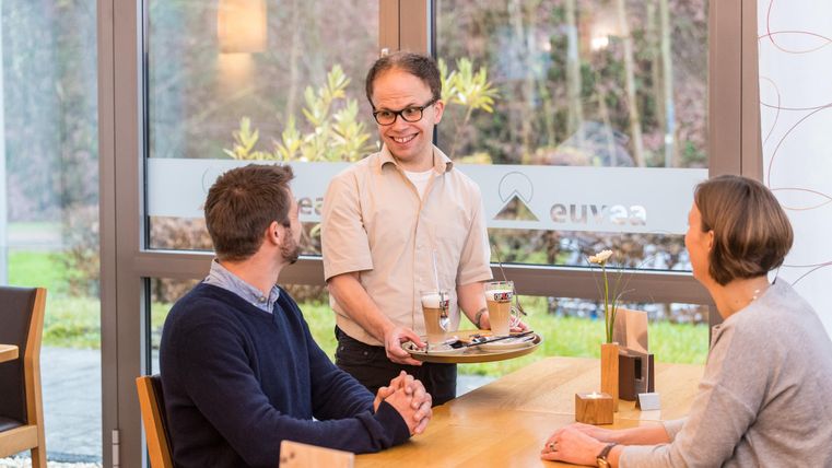 Een ober brengt drankjes naar een tafel met twee gasten in een modern café. De sfeer is vriendelijk en uitnodigend.