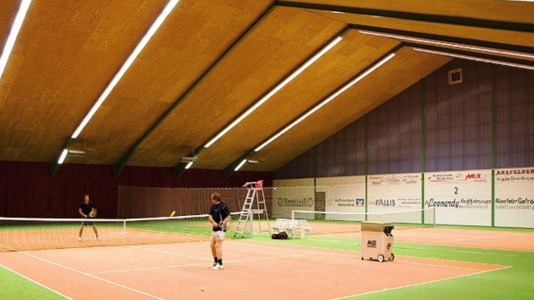 Eine moderne Tennishalle mit einem orangefarbenen Platz und grünem Teppich. Zwei Spieler trainieren im Innenbereich.