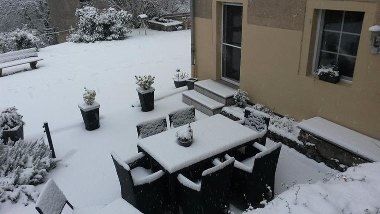 A snow-covered garden with a table and chairs covered in snow. In the background, a few plants and a house are visible.