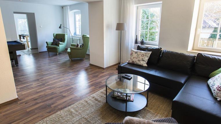 A modern living room with a cozy couch and a table. In the background, there are green armchairs and a bright room.