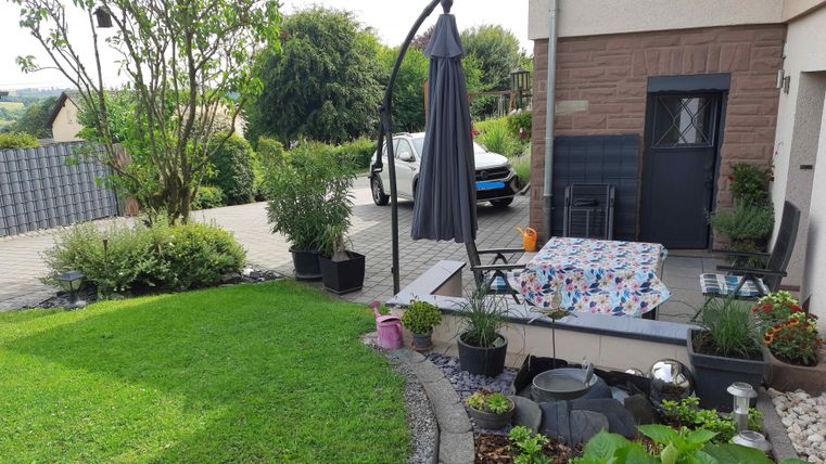 Ein schöner Garten mit Rasen, Pflanzen und einer Terrasse. Im Hintergrund steht ein Auto und ein paar Bäume sind sichtbar.