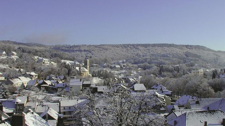 Eine verschneite Winterlandschaft mit einem kleinen Dorf und vielen Häusern. Der Himmel ist klar und blau, umgeben von bewaldeten Hügeln.