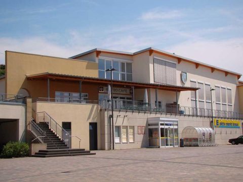 Ein modernes Gebäude mit einer großen Terrasse und einem Eingangsbereich. Vor dem Gebäude befinden sich Stufen und eine gepflasterte Fläche.