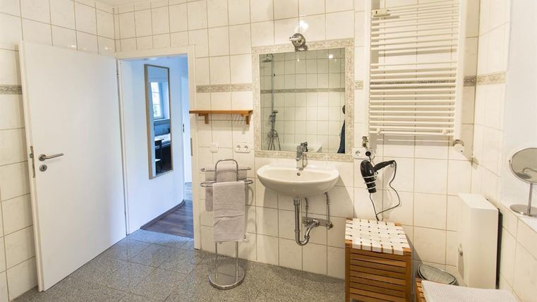 A modern bathroom with white tiles and a sink. There is a mirror, a shower, and a small wooden shelf.