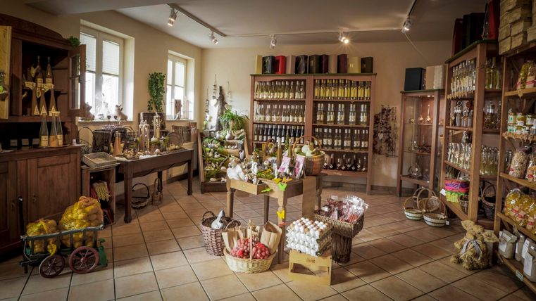 Ein gemütlicher Laden mit Regalen voller Vorräte und Genussmitteln. In der Mitte stehen Körbe und Tische mit verschiedenen Produkten.