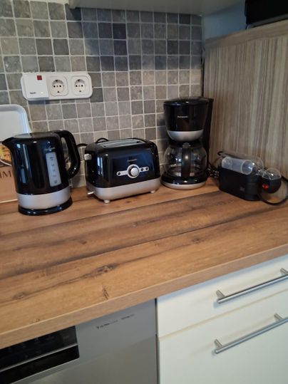 Un comptoir de cuisine moderne avec une bouilloire, un grille-pain, une cafetière et un robot culinaire. Les appareils sont en noir et en acier inoxydable et se trouvent sur une surface en bois.