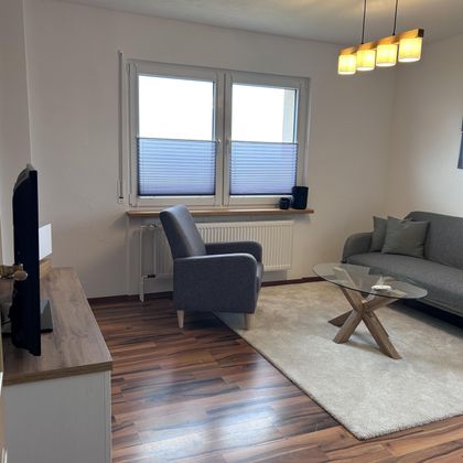 Een moderne woonkamer met een grijze bank en een fauteuil. Er is een houten vloer en een vierkante tafel van hout.