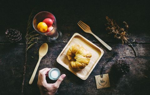 Une table rustique avec un plateau en bois sur lequel se trouvent une pâtisserie et une coupe en verre avec des fruits frais. Une main tient un petit verre tandis qu'une cuillère est posée à côté.