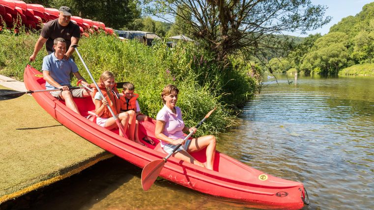 Een gezin peddelt in een rode kajak op een rustige water. Op de achtergrond zijn bomen en een zonnige lucht te zien.
