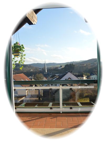 A view from a balcony overlooking a picturesque landscape with rooftops and hills in the background. The sky is clear and sunny.