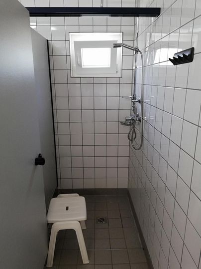 Een eenvoudige douchecabine met witte tegels en een raam. Een plastic zitje staat in de ruimte.