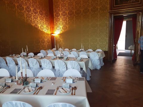 Ein eleganter Bankettsaal mit festlich gedeckten Tischen und Stühlen in Weiß. Die Wände sind mit goldenen Mustern dekoriert, und sanftes Licht schafft eine einladende Atmosphäre.