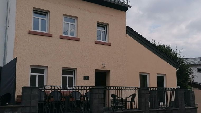 Ein modernes Haus mit einem hellen Außenanstrich und mehreren Fenstern. Vor dem Haus befindet sich eine kleine Terrasse mit Stühlen.