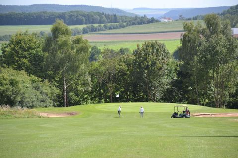 Eine weitläufige Golfanlage mit grünen Fairways und Bäumen im Hintergrund. Zwei Golfer sind auf dem Platz, während ein Golfwagen in der Nähe steht.