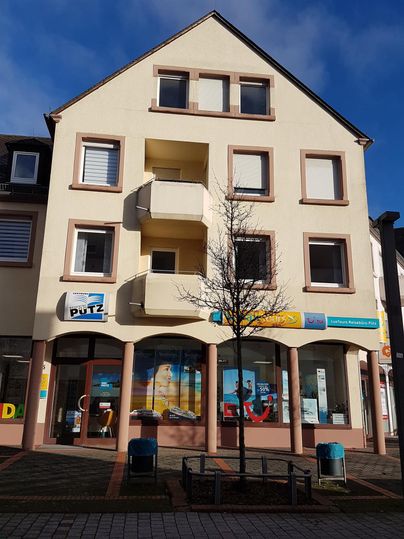 Ein modernes Gebäude mit mehreren Fenstern und Balkonen. Im Erdgeschoss sind Geschäfte mit bunten Schildern.