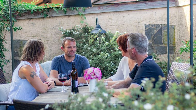 Eine gesellige Runde von vier Personen sitzt an einem Tisch im Freien. Sie genießen Getränke und unterhalten sich in einer freundlichen Atmosphäre.