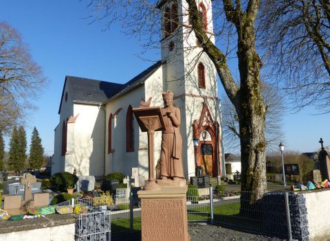 Eine Kirche mit einem hohen Turm und einer Statue im Vordergrund. Der Hintergrund zeigt einen Friedhof mit Bäumen und sonnigem Wetter.