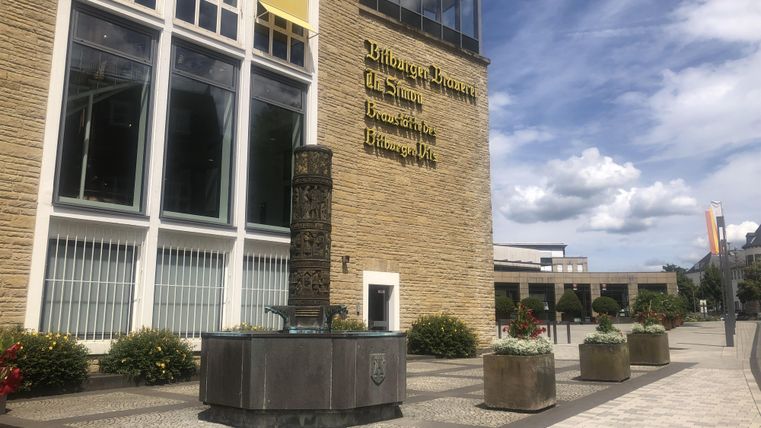 Ein modernes Gebäude mit einer großen Glasfassade. Vor dem Gebäude steht ein runder Brunnen, umgeben von Pflanzkübeln.