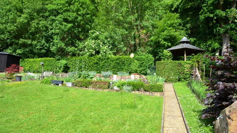 A beautiful garden with green grass and dense shrubbery. In the background, you can see a gazebo and beds with various plants.