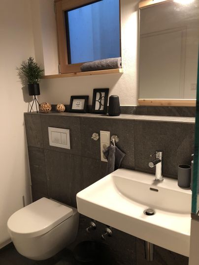 Ein modernes Badezimmer mit einer grau gefliesten Wand und einem eleganten Waschbecken. Ein großer Spiegel und eine kleine Pflanzendekoration sorgen für eine einladende Atmosphäre.