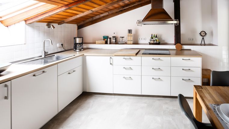 A modern kitchen with white cabinets and wood surfaces. The roof is slanted, and there are large windows that let in plenty of light.