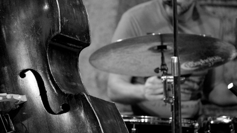 Ein Kontrabass steht im Vordergrund, während ein Schlagzeuger im Hintergrund spielt. Die Szene vermittelt eine lebendige Atmosphäre einer Musikaufführung.