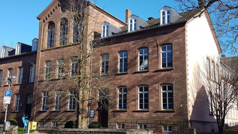 Ein historisches Gebäude mit roten Backsteinmauern und großen Fenstern. Es befindet sich in einer ruhigen Umgebung mit Bäumen und einem kleinen Platz davor.