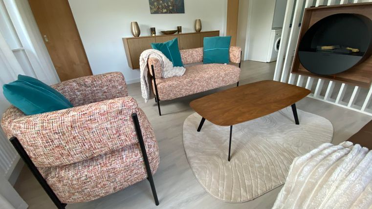 Ein modernes Wohnzimmer mit zwei gemütlichen Sesseln, einem Holztisch und einer hellen Farbpalette. Im Hintergrund sind dekorative Elemente und ein Teppich zu sehen.
