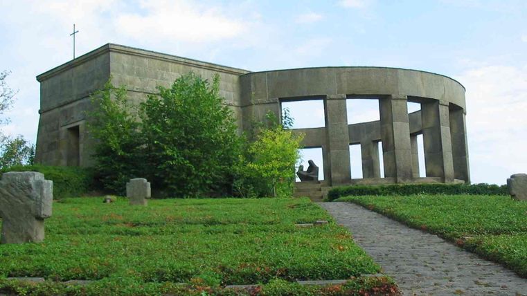 Ein monumentales Gebäude, umgeben von grünen Wiesen und Bäumen. Der Eingang ist von Säulen umgeben und der Himmel ist leicht bewölkt.
