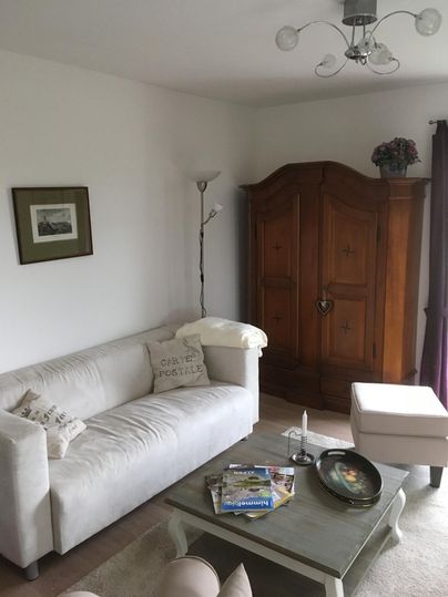 Ein gemütliches Wohnzimmer mit einem weißen Sofa und einem großen Holzschrank. Auf dem Tisch liegen einige Zeitschriften und ein dekorativer Teller.